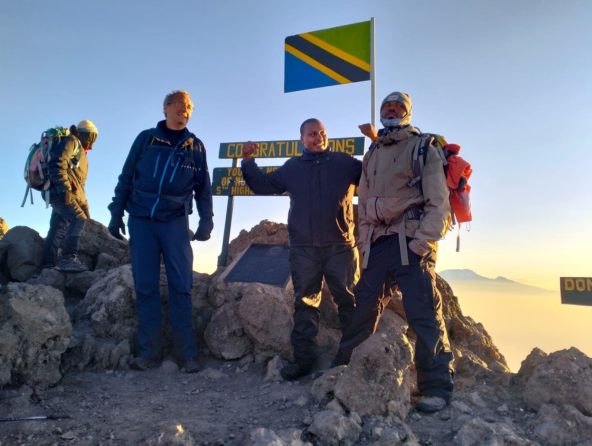 Trust Tours climbers and guide celebrating with the Tanzanian flag at a summit sign